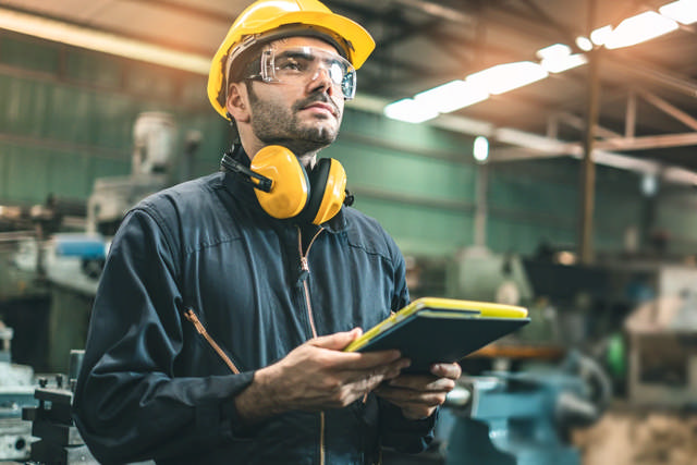 Mann in industrieller Umgebung mit Helm, Schutzbrille und Gehörschutz, hält ein Tablet in der Hand – Beispiel für moderne Sicherheitsausrüstung