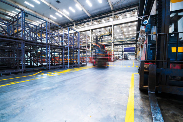 Industriehalle mit gelben Bodenmarkierungsbändern, die sichere Verkehrswege und Lagerbereiche definieren, während ein Gabelstapler in Bewegung ist.