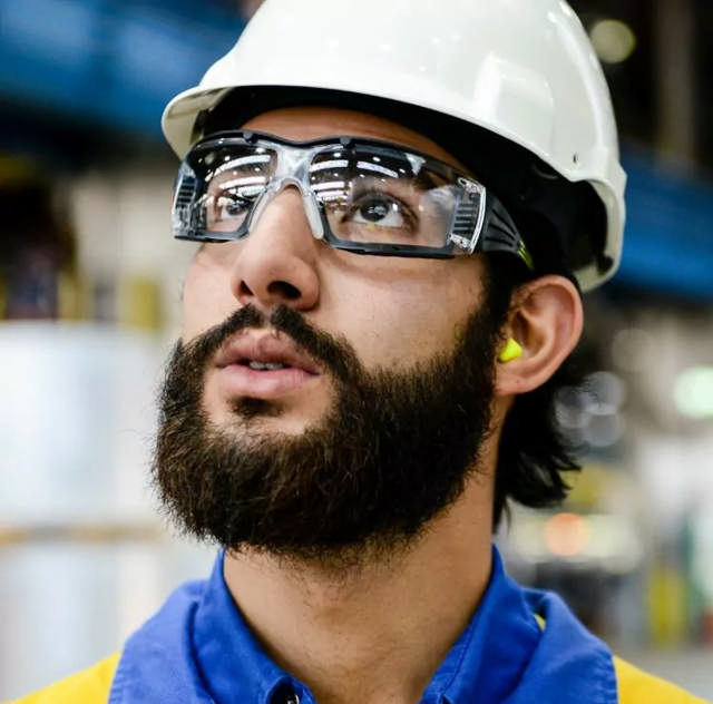 Mann mit Schutzbrille, Helm und Gehörschutzstöpseln, konzentriert bei der Arbeit in einer industriellen Umgebung.